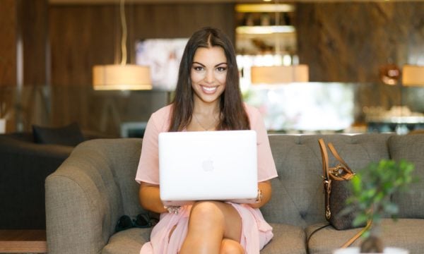 A woman sitting on a sofa, holding a laptop, looking at and smiling into the camera.
